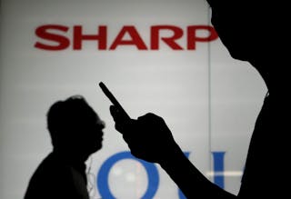 A man using his mobile phone walks past a logo of Sharp Corp outside an electronics shop in Tokyo