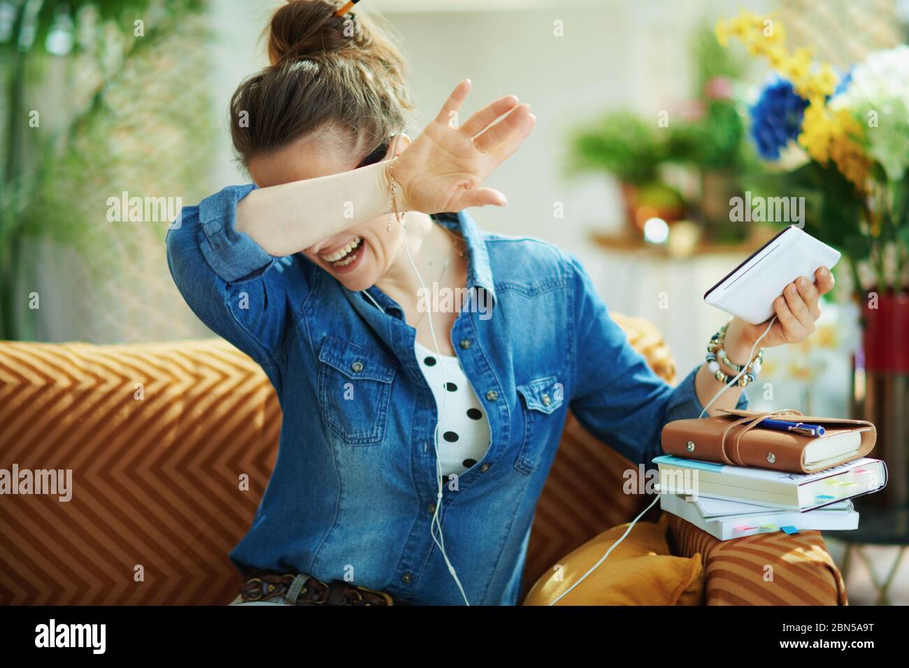 happy young housewife in jeans shirt with white headphones and tablet PC rejoicing while study online in the modern living room in sunny day. Stock Photo