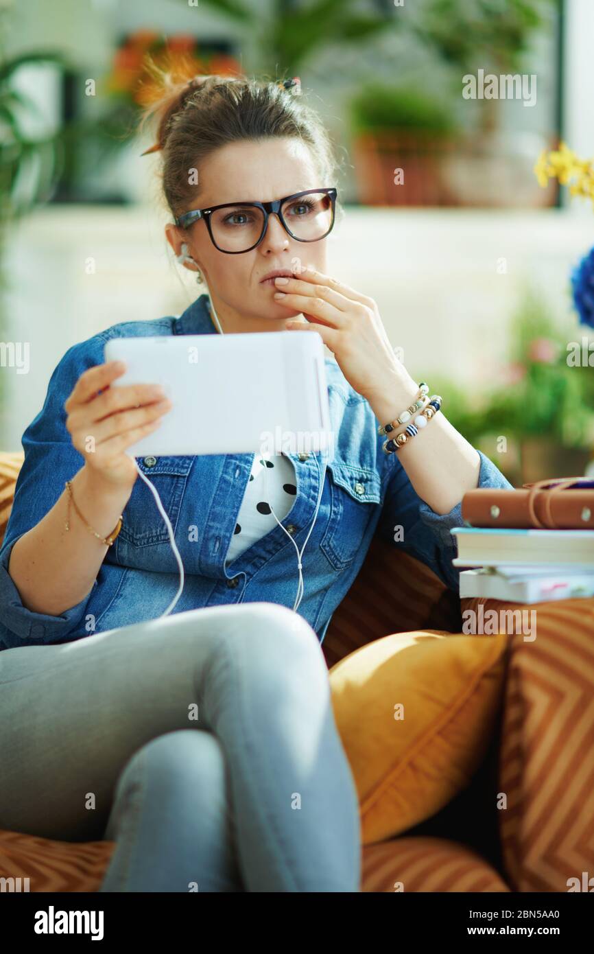 modern housewife in jeans shirt with white headphones and tablet PC study online in the modern living room in sunny day. Stock Photo