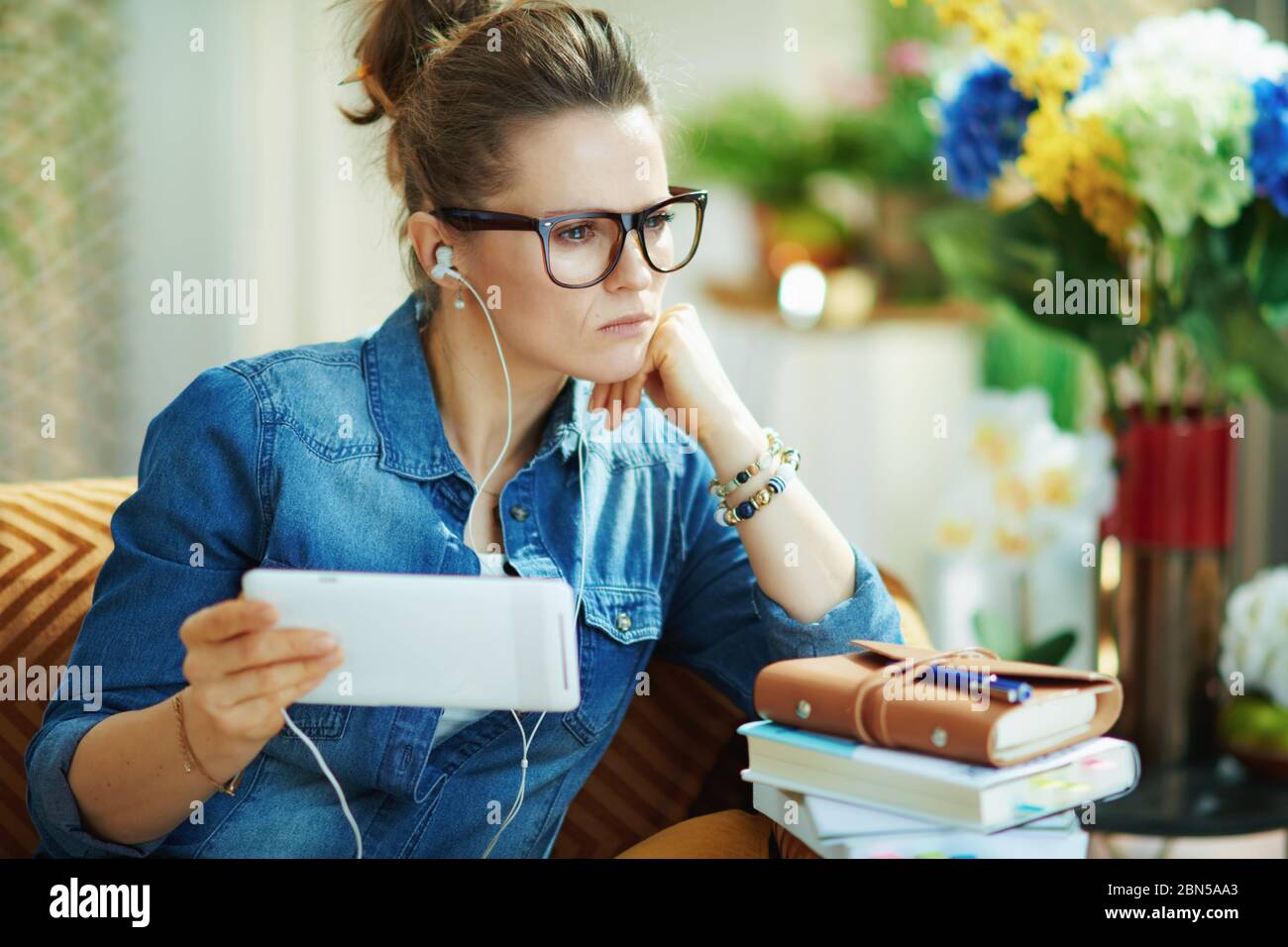 pensive stylish female in jeans shirt with white headphones and tablet PC study online in the modern living room in sunny day. Stock Photo