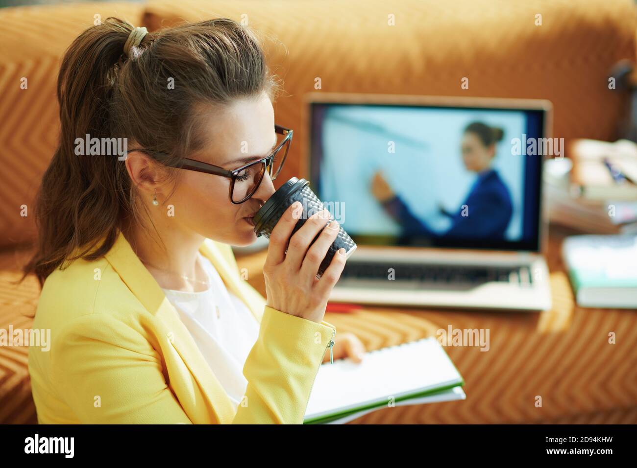 young housewife in yellow jacket in the modern house in sunny day drinking coffee and study online on a laptop. Stock Photo