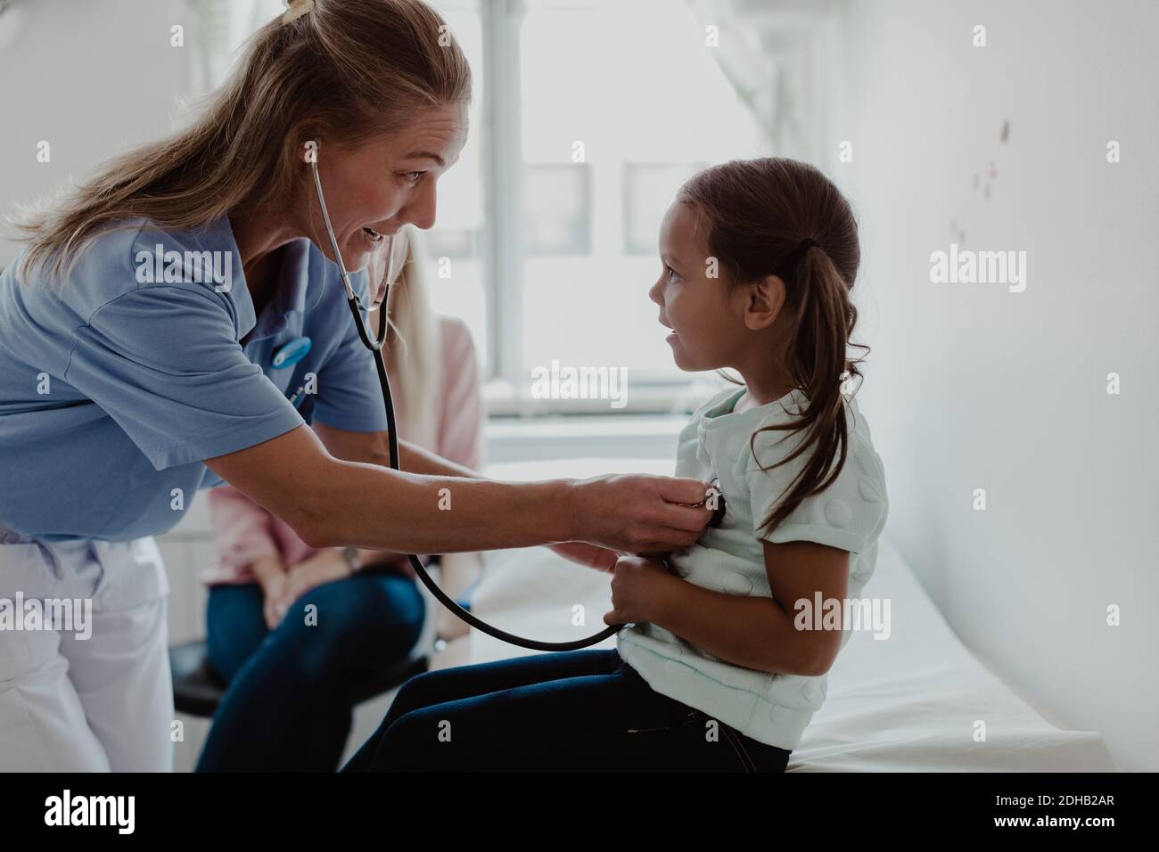 Female pediatrician examining girl's heartbeat through stethoscope while mother sitting in background at clinic Stock Photo