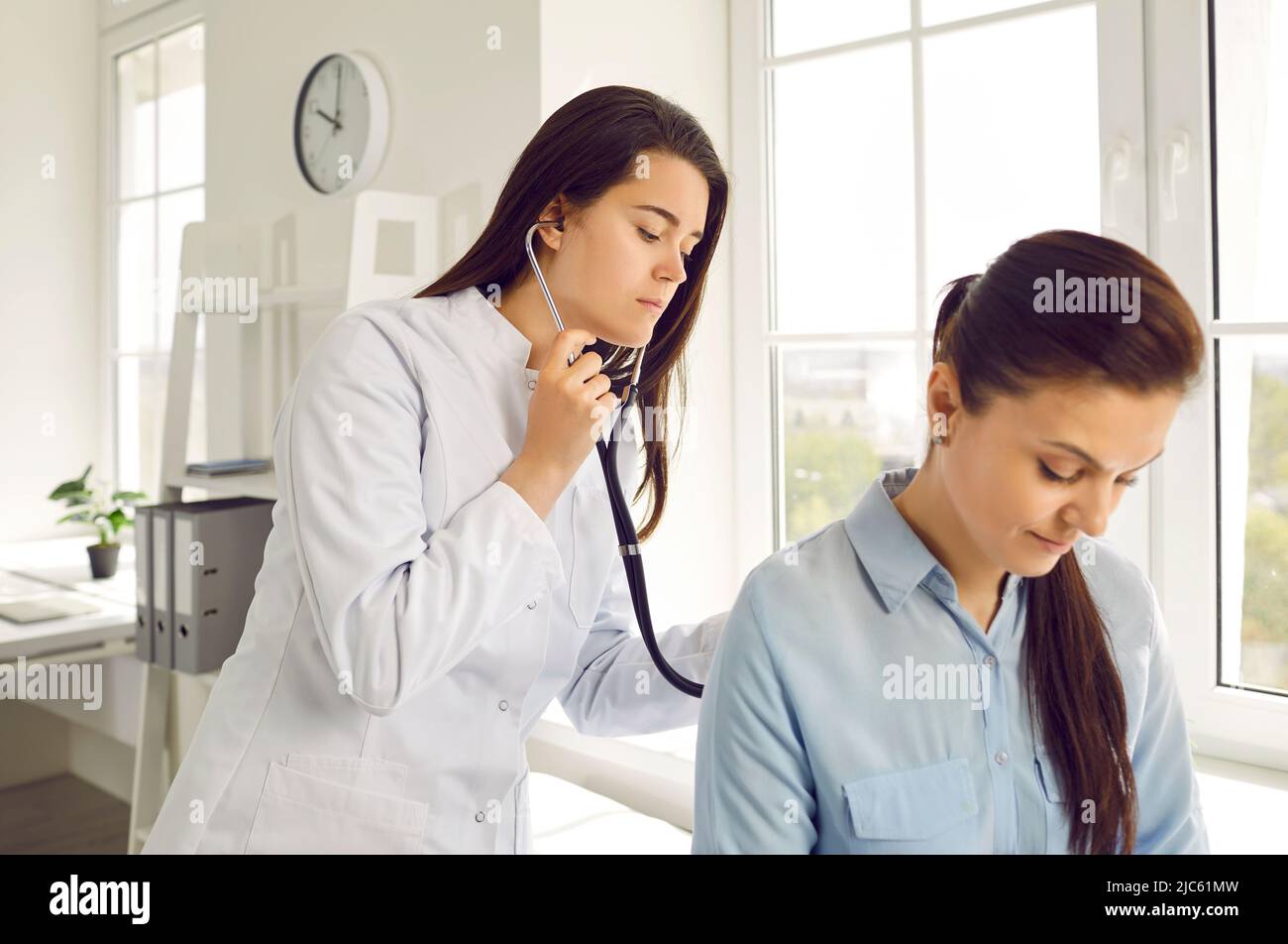 Young female doctor checks woman's breathing and heartbeat at appointment at medical clinic. Stock Photo