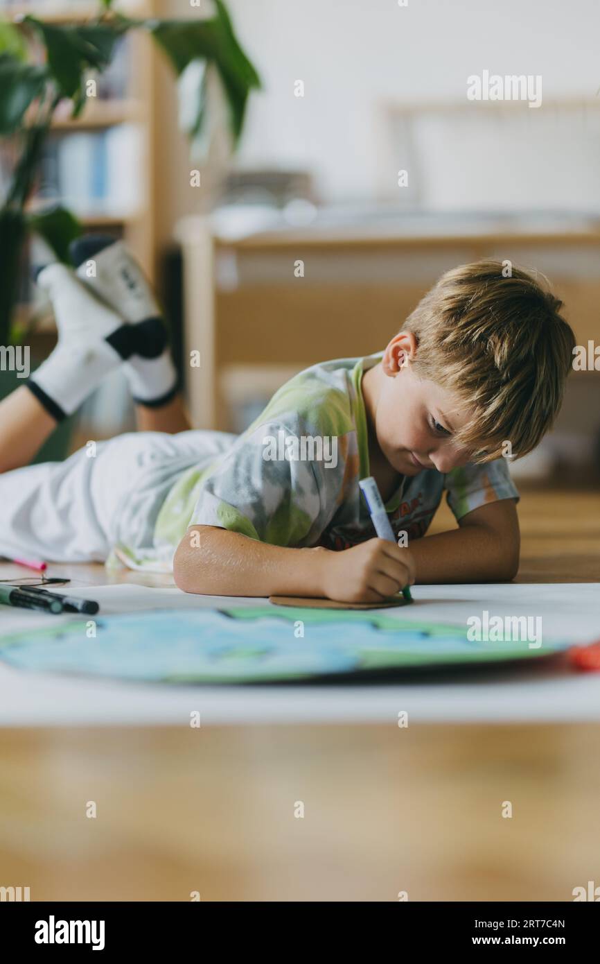 Boy lying on stomach and painting at home with watercolors and tempera paints, creating a model of planet Earth. Stock Photo