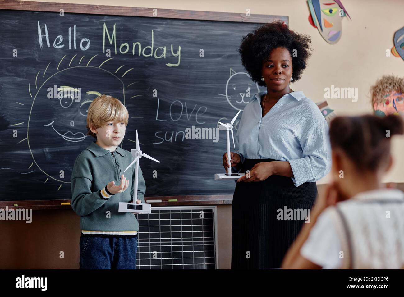 Shot of teacher of Black ethnicity and primary school boy answering at blackboard showing small wind turbine with solar panel to class Stock Photo