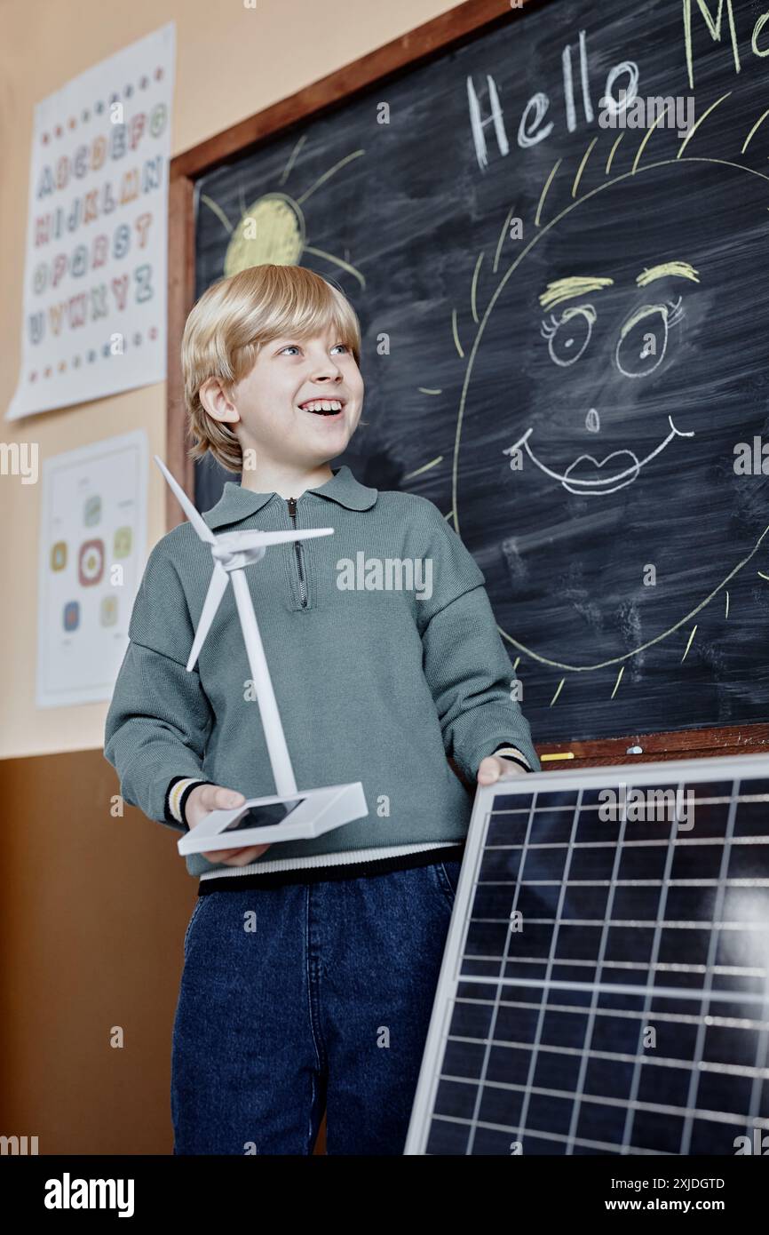 Vertical shot of cheerful primary learner presenting green energy project holding small solar powered wind turbine and big solar panel showing devices to class Stock Photo