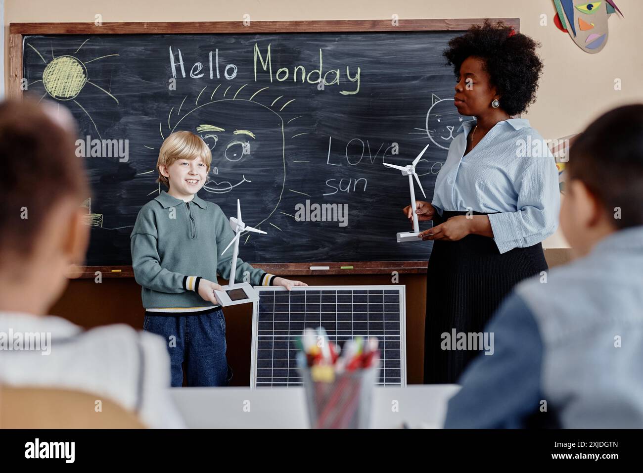 Smiling young boy of primary school showing big solar panel and small wind turbine to classmates answering at blackboard with funny sun drawings Stock Photo