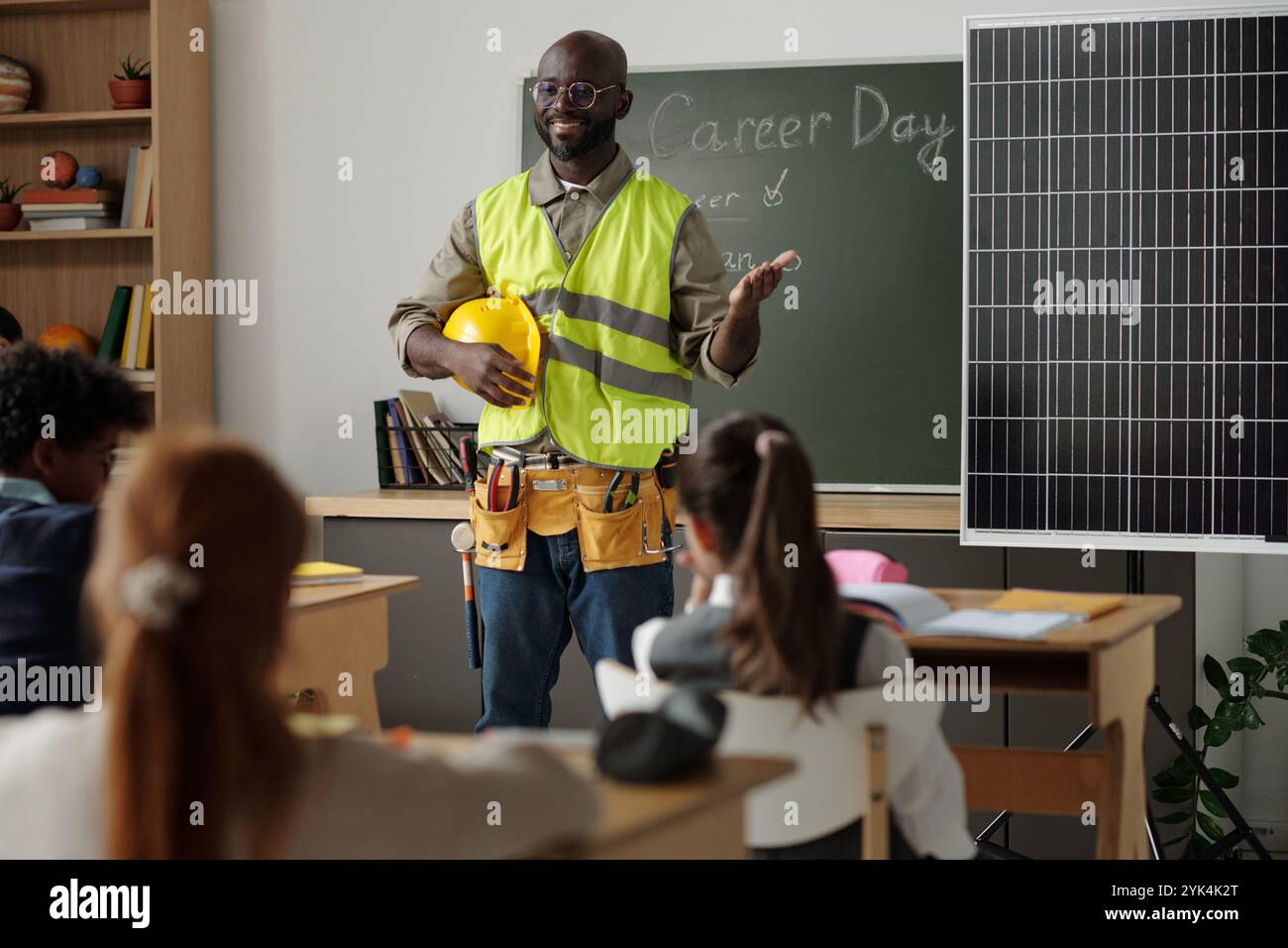 Young smiling African American engineer in reflective vest making presentation of solar panel model to schoolkids Stock Photo