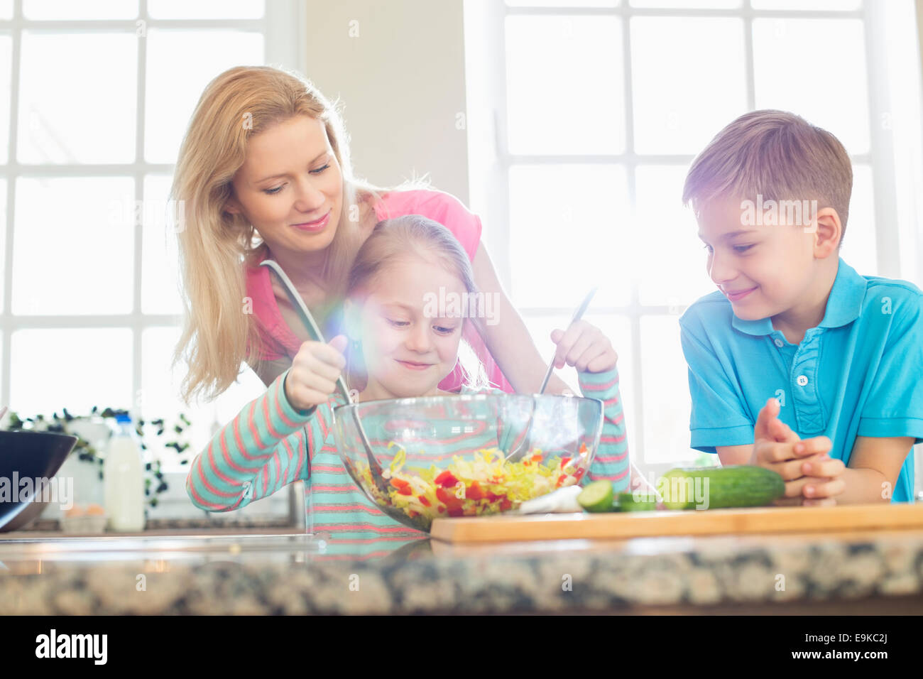 Family looking at girl mixing salad in kitchen Stock Photo