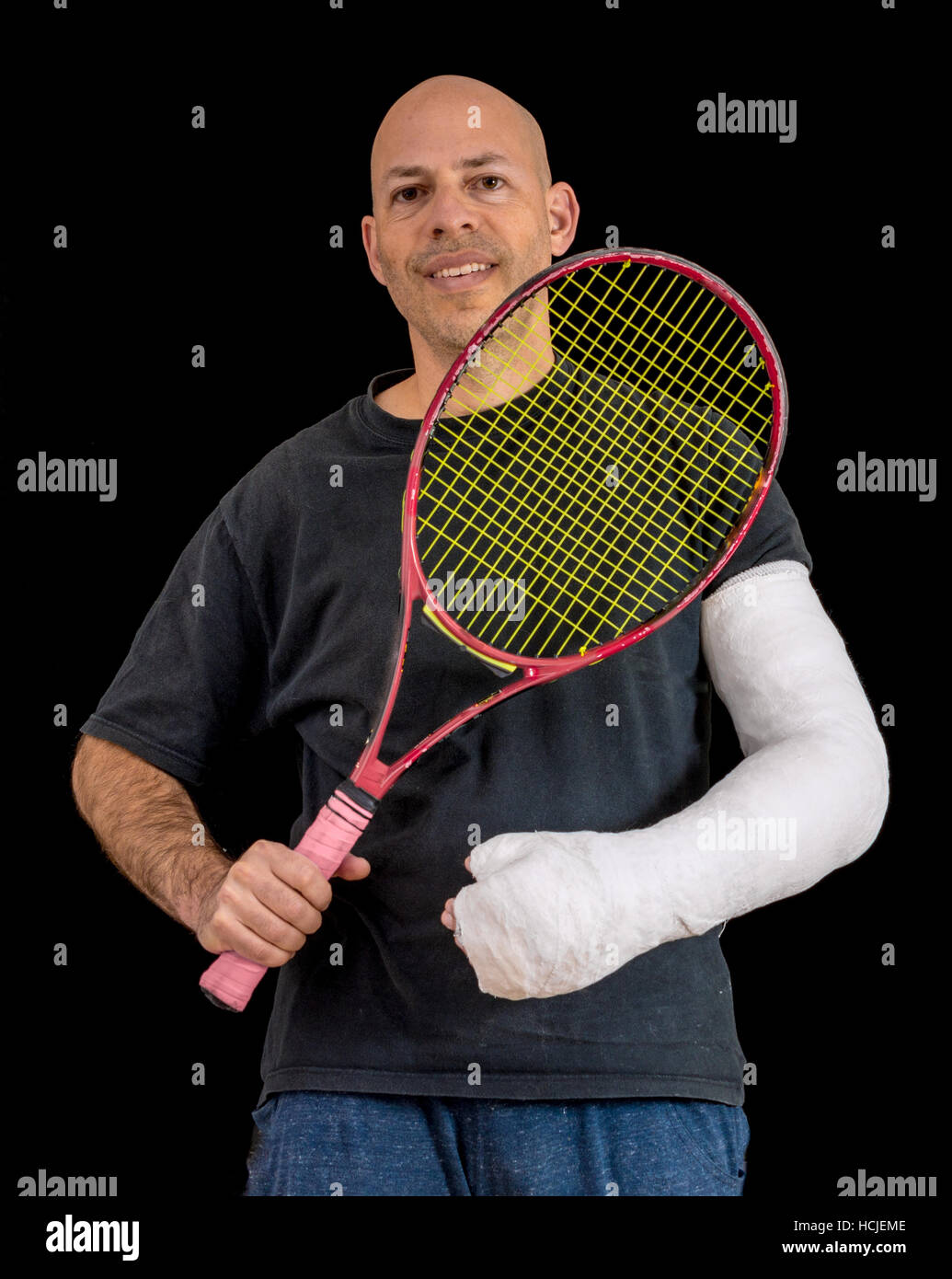 Young man holding a tennis racket after breaking his wrist in a tennis game, sporting a bright white arm cast, isolated on black Stock Photo