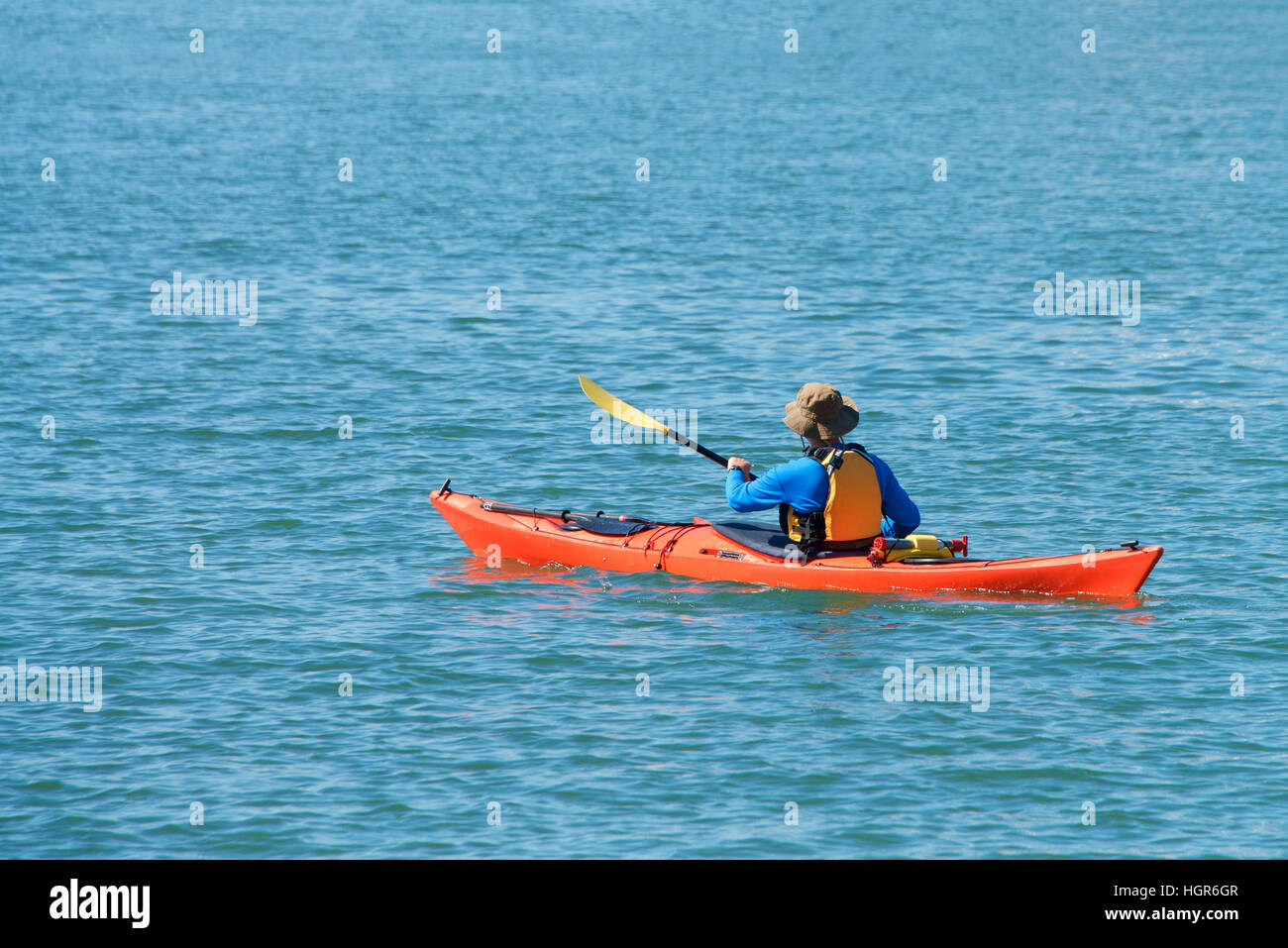 person paddling in an orange kayak wearing life jacket surrounded by water, facing away from camera traveling right to left across the frame. Stock Photo