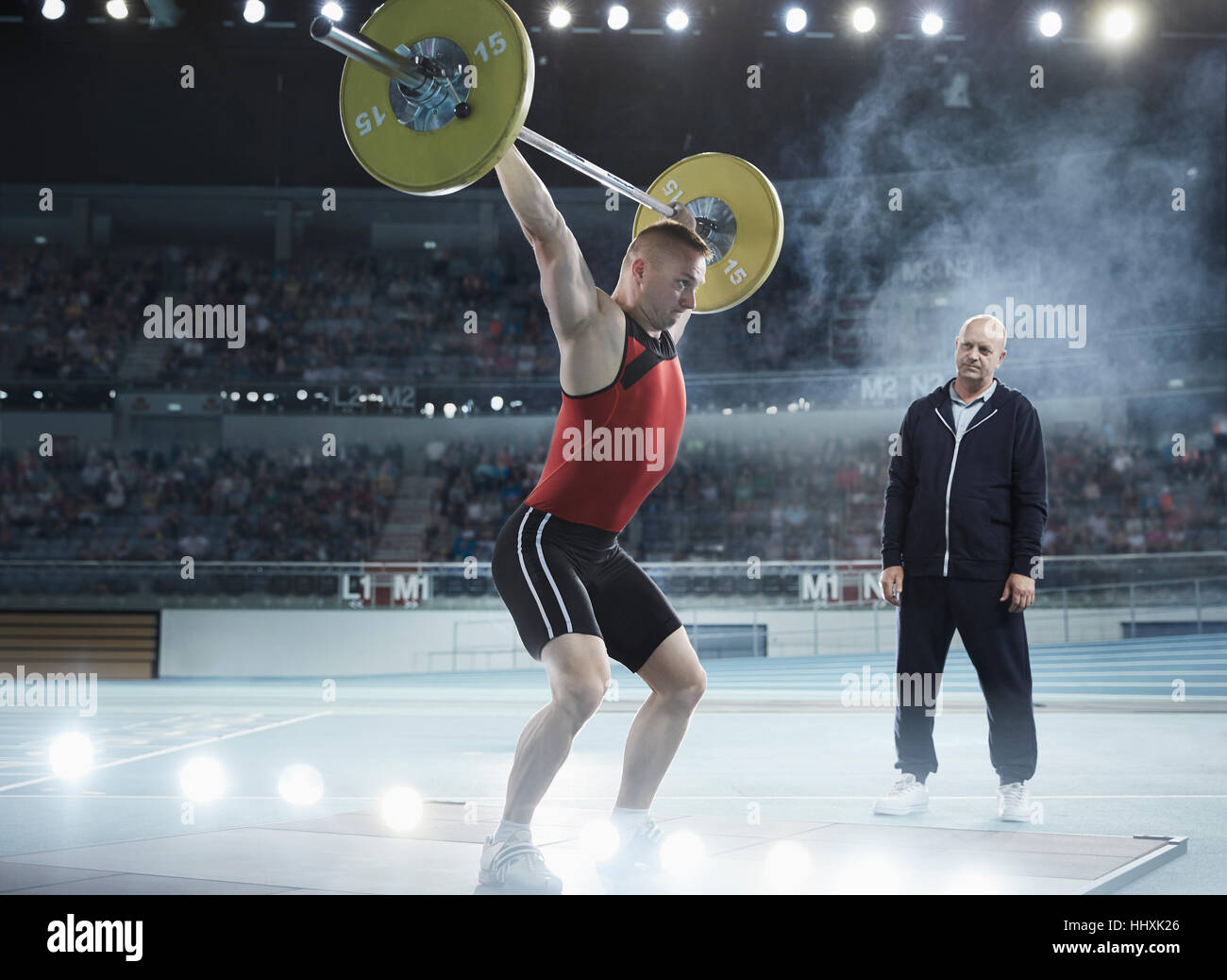 Coach watching male weightlifter squatting barbell overhead in arena Stock Photo