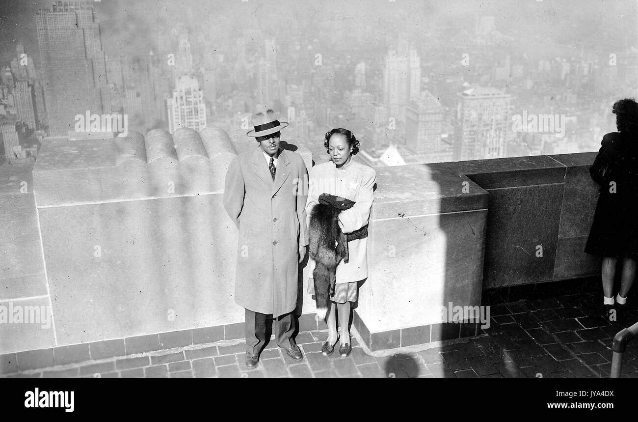 African-American couple, a man and a woman, standing together near a railing at the top of the Empire State Building, on an observation deck, viewed from above, the skyline of New York visible in the background, the man wearing a trench coat and formal hat, the woman wearing a shirt and holding a fur coat, 1946. Stock Photo