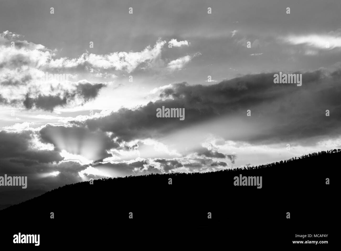 Sunset over a trees line on a hill, with sun rays coming out through some clouds Stock Photo