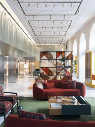 Two red sofas with red cushions and two red patterned chairs surround a glass topped table. Behind, a four-by-four display square shelf unit. Additional furniture behind.