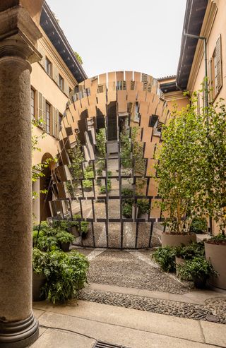 A courtyard featuring a large tower of reflective square mirrors surrounded by plant pots on the floor.