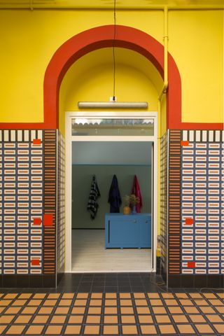 A tiled door way which leads into another room with a low blue cabinet and three coats hanging on a wall. Above the door frame the wall has been painted in yellow with a red border.