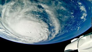 The bulbous blue orb of Earth as seen from above. On its face, on the left, a giant white swirl of this cloud, a hurricane. On the bottom right, within the sliver of black space beneath the Earth, a white spacecraft is partially seen.