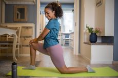 Woman performs hip flexor stretch at home. She is in a half kneeling position with her hands on her front knee. 