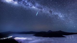 The Milky Way stretches across the sky and a bright meteor streaks across the center. Below is the top of Mount Bromo surrounded by clouds.