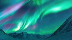 Green and purple northern lights appear over the sky above snowy mountains in Alaska, USA