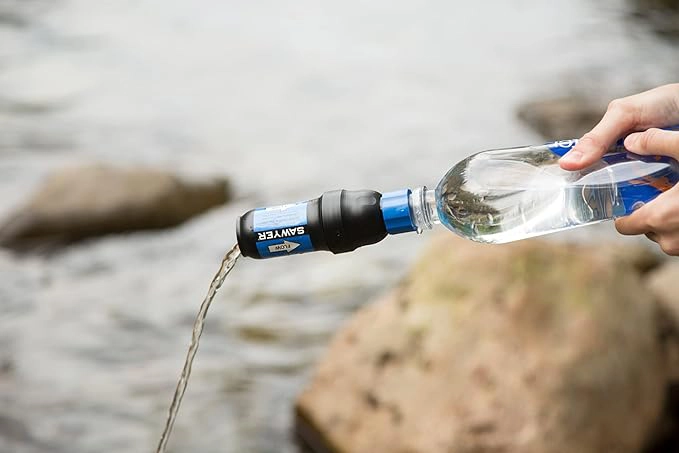 A Sawyer cleaning coupling attached to a Smartwater bottle and Sawyer squeeze filter backflushing into a body of water