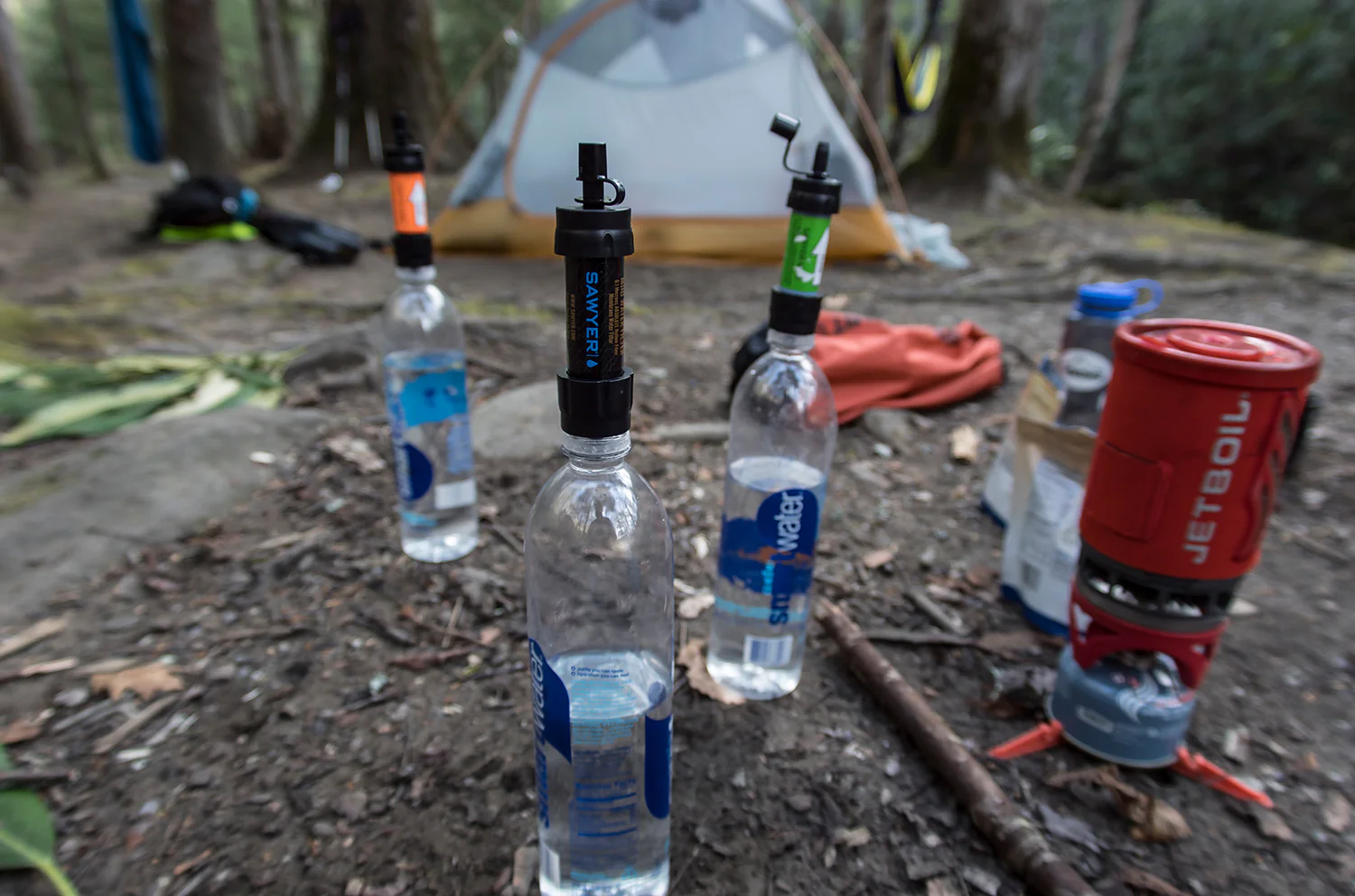 Three multi-colored Sawyer mini water filters attached to three Smart water bottles in front of a tent in the forest