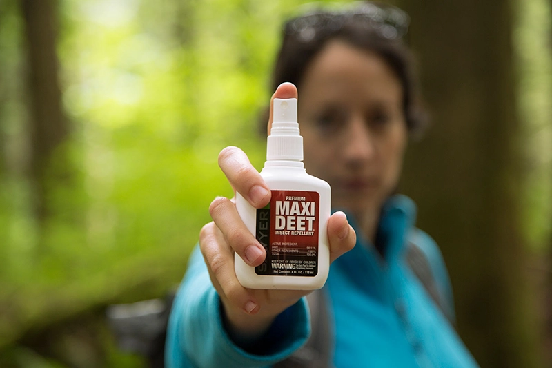 A person holding a bottle of Sawyer premium maxi deet insect repellent towards the camera in the forest