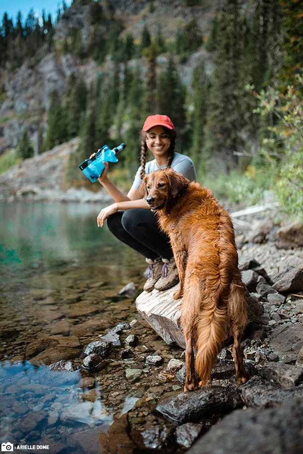 A woman holding a Sawyer squeeze water pouch attached to a mini water filter next to a dog by the lake