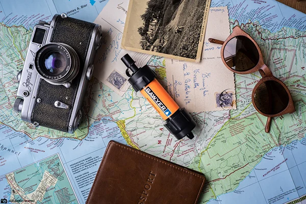 An orange Sawyer mini water filter sitting on top of a map with photos, a camera, sunglasses and a passport book