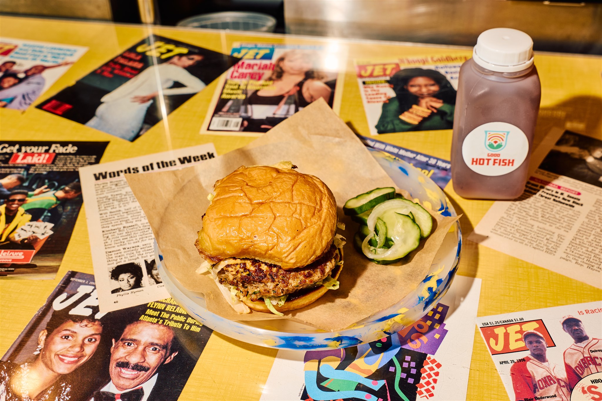 A fish sandwich with a side of pickles on a plate on top of a counterop covered with the pages of “Jet” magazines.