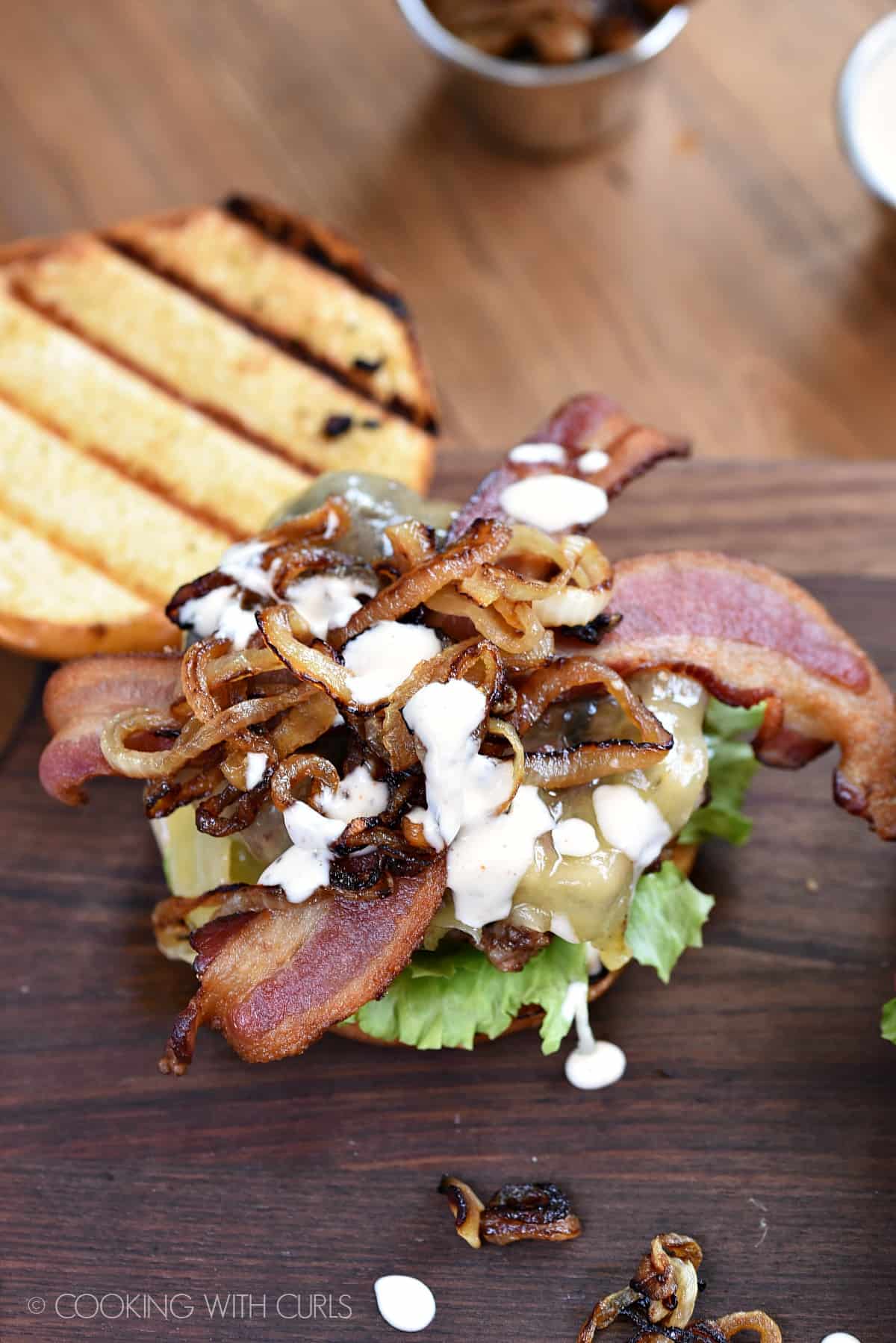 Looking down on an open faced Grilled Double Bacon Burger topped with caramelized onions and white barbecue sauce with a grilled bun sitting on the side.