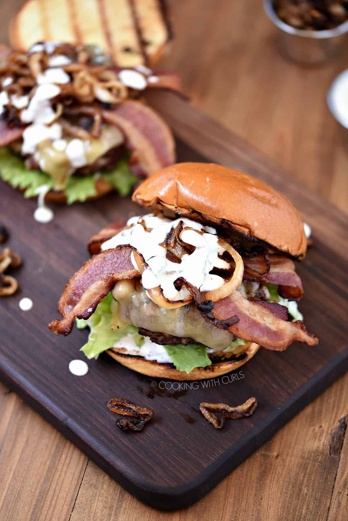 Looking down on two Grilled Double Bacon Burgers topped with caramelized onions and white barbecue sauce sitting on a wood board.