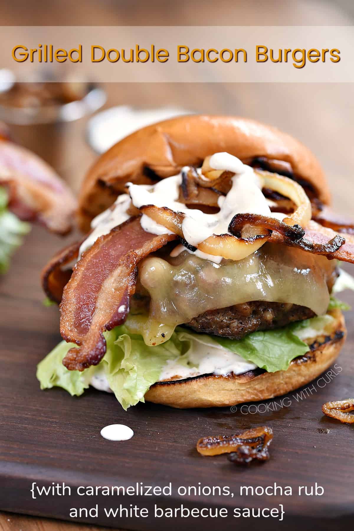 Grilled Double Bacon Burgers on grilled buns, topped with lettuce, tomato, caramelized onions and white barbecue sauce sitting on a wood board with a title graphic across the top.