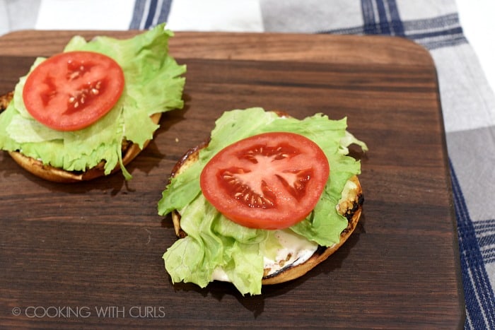 Lettuce and a tomato slice on top of the grilled burger buns sitting on a walnut cutting board.