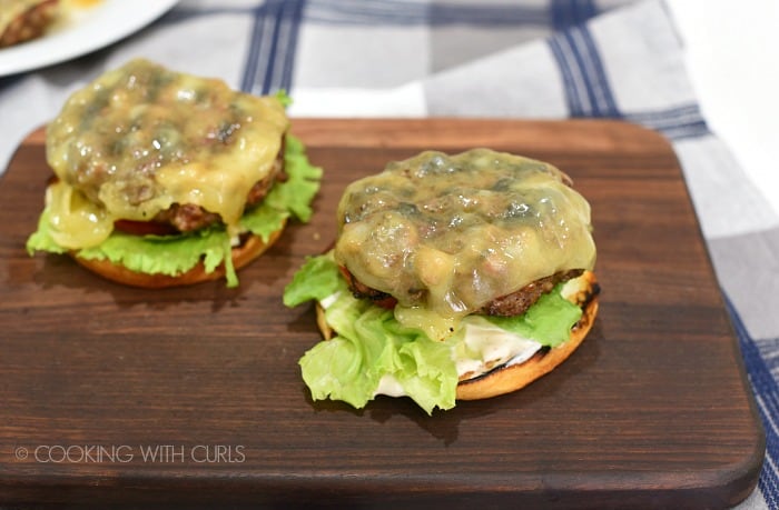 Two buns topped with white barbecue sauce, lettuce, tomato and grilled cheeseburger patties sitting on a walnut cutting board.