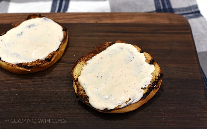 Two, grilled burger buns topped with white barbecue sauce sitting on a walnut cutting board.