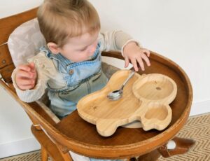 A young child sat in a wooden high chair holding a spoon in front of a wooden rabbit shaped plate