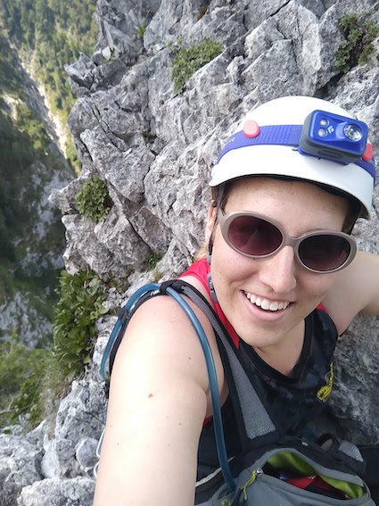 Mirah halfway up a the Drachenwand klettersteig in Austria. She is wearing a helmet and sunglasses and has a backpack with hydration system slung over her shoulder.