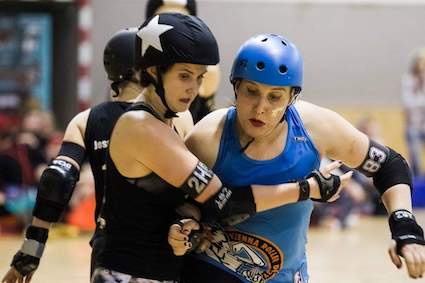 Mirah wearing protective equipment and a Vienna Roller Derby jersey blocking the opposing jammer with her hips. Mirah has a look of concentration on her face.