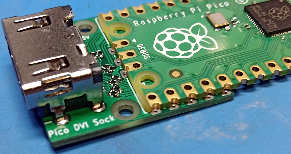 A Pico board rests lightly on top of a sock. The holes on the two boards are lined up so that you can see through both boards at each of the 10 IO pads on the board edges.