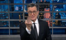 A man in a suit stands on a talk show stage holding a blurry middle finger up at the camera.