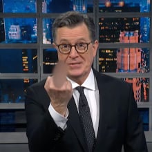 A man in a suit stands on a talk show stage holding a blurry middle finger up at the camera.