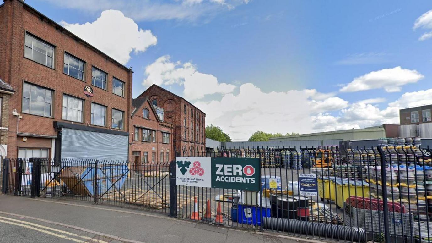 A large red brick building is on the left with a small plaque that reads Banks Brewery. There is a huge yard at the front with hundreds of metal beer kegs piled on top of each other, behind a black metal fence.