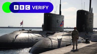 Russian submarines docked in Tartous in 2019. An armed soldier stands next to the vessels on a dock. 