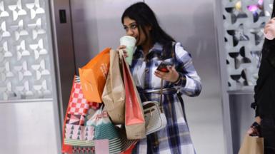 A woman sips coffee in a store while holding multiple shopping bags on one arm