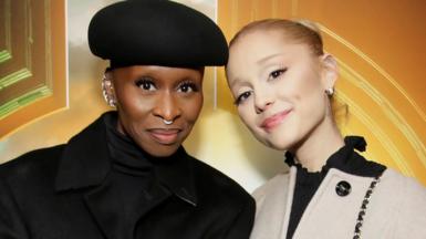 Actresses Cynthia Erivo and Ariana Grande stand next to each other, their heads leaning in towards each other as they smile at the camera. Erivo is dressed in all black, and wears a hat. Grande wears a beige coat over a black top, with her hair tied back.