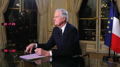 Michel Barnier, who has grey combed over hair and wears a black suit with a white shirt and a black tie, looks towards a camera during a TV interview at the Hotel Matignon in Paris 