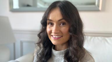 A profile of Hannah Newby wearing a light grey woolly jumper and long, curly dark hair sitting on a sofa in front of a mirror.