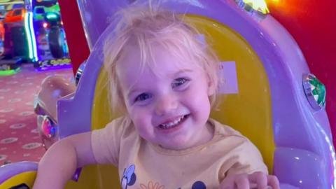 Isabella Jonas-Wheildon, a young blonde toddler wearing a yellow t-shirt smiling at the camera. She is sitting on a yellow and purple chair at an arcade.
